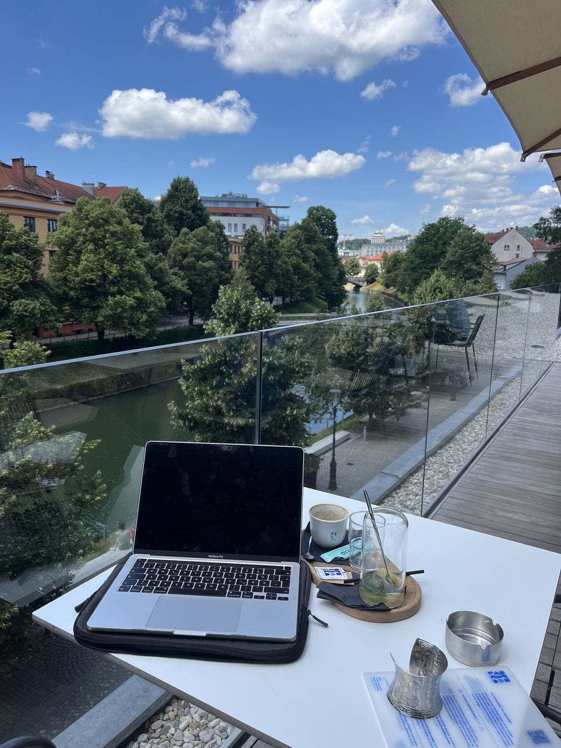 Odprt MacBook na beli mizi v kavarni nad Ljubljanico, ledena kava, strehe starega mesta.