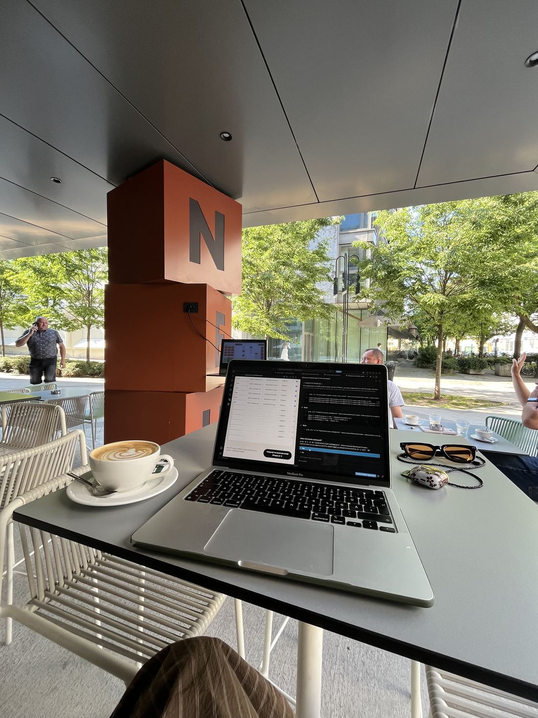 MacBook in latte na mizi zunanje kavarne, drevesa in oranžen steber v ozadju.